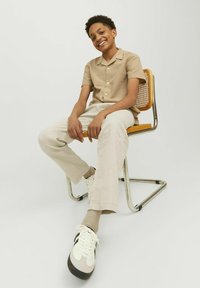 Lichtbruine shirt met korte mouwen van textuurstof, crèmekleurige broek en witte sneakers met zwarte accenten, zittend op een metalen stoel.