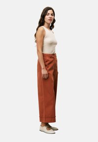 Débardeur côtelé crème, pantalon large rouille avec ourlets retroussés et poches latérales, et chaussures à lacets de couleur claire.