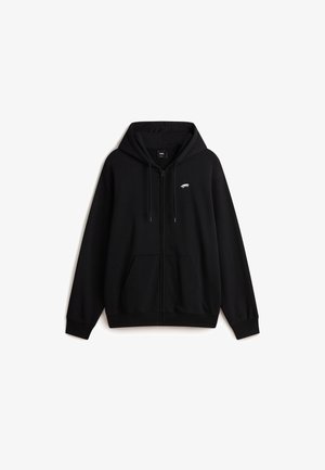 Persona que lleva una sudadera negra con cremallera, pantalones cortos vaqueros azul claro, calcetines blancos de Vans y zapatillas Vans azules con suelas marrones.