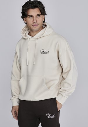 Sudadera beige con un bolsillo frontal y cordón, con un logo negro en el lado izquierdo del pecho. Combinada con pantalones marrón oscuro con acento de logo.
