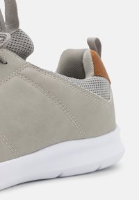 Lys grå sneaker med semsket ytterside, mesh-tunge, hvit såle og en brun skinnaccent nær hælen. Synlig sømdetalj.