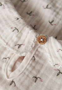 Blød hvidt stof med små sorte og lyserøde blomsterprints, bølget blondekrave og en brun træknap, der fastgør en nøglehulåbning.