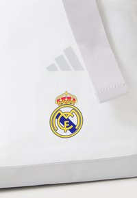 White sports bag with gray accents, featuring the Adidas logo and a colored Real Madrid crest with a crown. Smooth texture.
