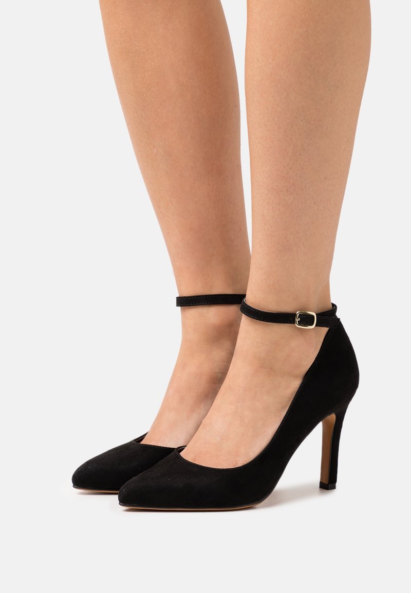 Escarpins à bout pointu en daim noir avec brides à la cheville et boucles dorées portés sur des jambes nues sur un fond uni.