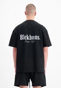Zwarte katoenen T-shirt met grote witte geborduurde tekst op de achterkant: "Blckbnns Design Dept." Korte mouwen en een ontspannen pasvorm.