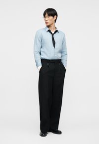 Άνδρας που φορά ανοιχτό μπλε πουκάμισο με κουμπιά, μαύρο φαρδύ παντελόνι, χαλαρή μαύρη γραβάτα, μαύρα παπούτσια και γυαλιά, στέκεται με τα χέρια στις τσέπες.