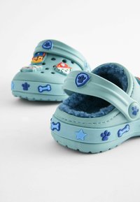 Saboti de culoare bleu deschis, cu căptușeală texturată din fleece, având accente colorate în formă de personaje 3D și animale, precum și modele decorative cu oase și stele.