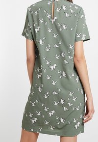 Kurzärmliges, locker sitzendes, grünes Kleid mit weißem Vogelprint. Öffnungsdetail am Rücken. Leichter Stoff mit glatter Textur.