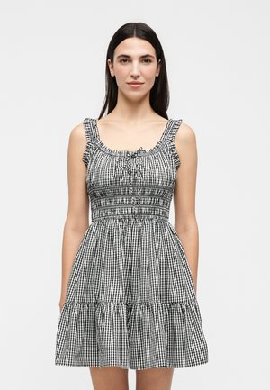 Donna con capelli neri e lisci che indossa un vestito senza maniche a quadretti bianchi e neri con spalline arricciate e una gonna a balze, in piedi davanti a uno sfondo semplice.