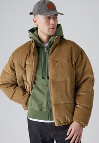 Brun kordfløjl puffjakke med ribbet tekstur, der har en grøn zip-up hætte trøje under. Inkluderer en grå kasket med et logo mærke.