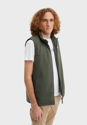 Olijfgroene vest gemaakt van lichtgewicht stof, met een hoge kraag, een ritssluiting aan de voorkant en twee zijzakken, gecombineerd met een wit T-shirt.