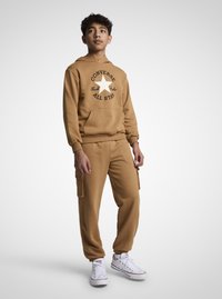 Hanorac maro din bumbac cu o stea albă și text negru, asortat cu pantaloni de trening cu buzunare laterale și manșete elastice. Adidași albi.