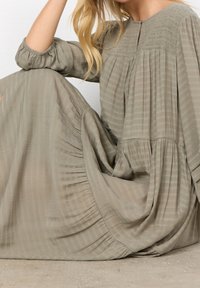 Maxi-jurk van licht olijfgroen stof met een textuur en geplooid ontwerp. Beschikt over een ronde halslijn en opgeblazen mouwen. Minimalistische esthetiek.