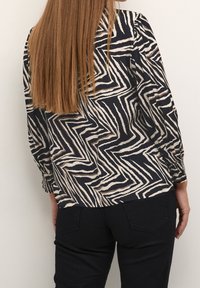 Blusa a maniche lunghe con un motivo a zigzag nero e crema, realizzata in tessuto leggero e con polsini elasticizzati. Abbinata a pantaloni neri.