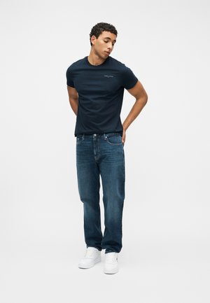 Mann står med hendene på ryggen, iført en marineblå Tommy Jeans T-skjorte, blå jeans og hvite sneakers mot en enkel hvit bakgrunn.