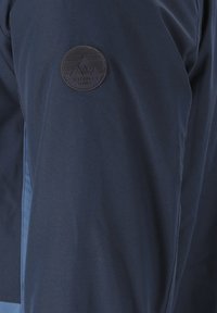 Marineblå jakke med en glat tekstur, prydet med et cirkulært, præget logo på den øverste del af armen, der står "WHISTLER DENMARK."