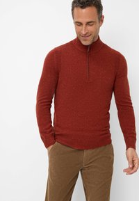 Roter Strickpullover mit hohem Kragen und Viertelreißverschluss, der eine strukturierte Optik aufweist, kombiniert mit braunen Cordhosen.