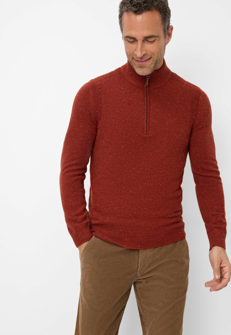Roter Strickpullover mit hohem Kragen und Viertelreißverschluss, der eine strukturierte Optik aufweist, kombiniert mit braunen Cordhosen.