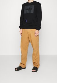 Sudadera negra con el logo "HUGO", pantalones de algodón beige y sandalias negras de tipo deslizante. Diseño sencillo, corte relajado y texturas suaves.