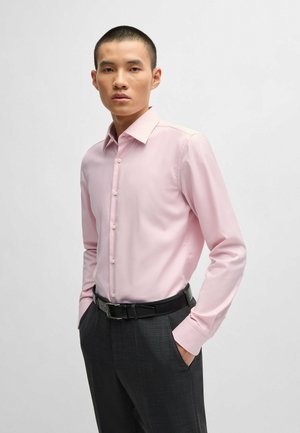 Chemise rose clair à manches longues avec un col standard et une fermeture à bouton, associée à un pantalon gris foncé et une ceinture noire. Texture lisse.
