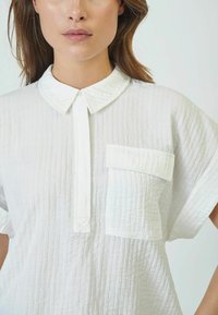 Camicia bianca realizzata in tessuto texturizzato, con colletto, maniche corte e taschino sul petto. Design semplice e minimalista.