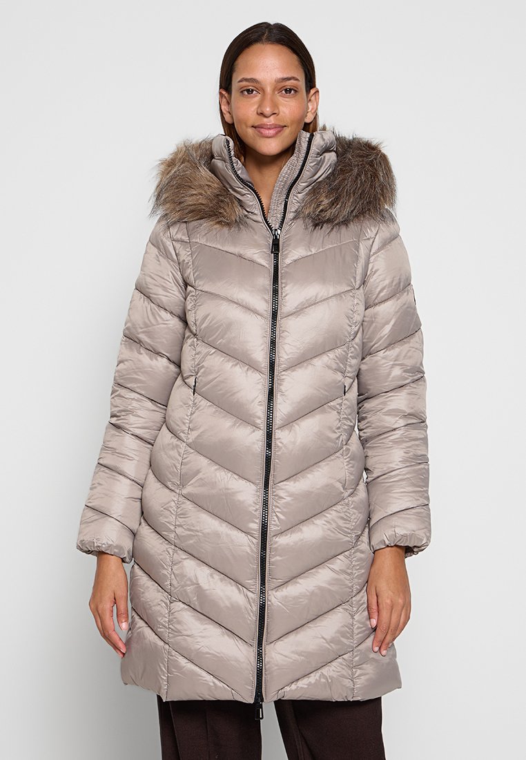 Donna che indossa un cappotto imbottito beige lungo fino al ginocchio, con cappuccio bordato in pelliccia, zip frontale e motivo a zigzag, in piedi davanti a uno sfondo semplice.