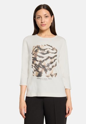 Woman with straight brown hair wearing a white long-sleeve shirt with an abstract tiger print and black pants standing against a white background.