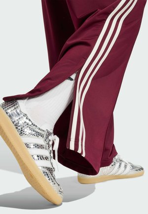 Nohy v bordových teplácích s bílými pruhy a zipy u kotníků, bílé ponožky a stříbrné texturované tenisky s gumovou podrážkou.