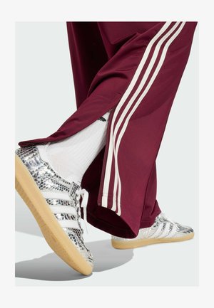 Piernas con pantalones deportivos burdeos con rayas blancas y cremalleras cerca de los tobillos, calcetines blancos y zapatillas plateadas texturizadas con suelas de goma.