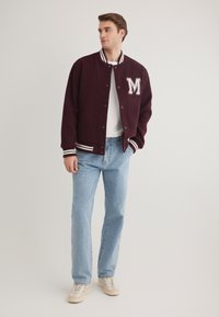 Bordeauxfarbene Collegejacke mit weißen Streifen an den Bündchen und dem Kragen, versehen mit einem auffälligen "M"-Patch. Getragen über einem weißen T-Shirt und hellen Jeans.
