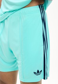 Ανοιχτό μπλε αθλητικό σορτς με μαύρο λογότυπο Adidas trefoil και τρεις μαύρες κάθετες ρίγες στο πλάι, φορεμένο από ένα άτομο.