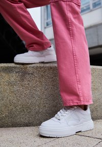 Vita sneakers med texturerade paneler, kraftiga plattformsålar och snörning fram, i kombination med rosa bomullsbukser. Står på en stenig yta.