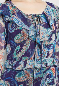 Květinové šaty v purpurové barvě, s volánkovým výstřihem a manžetami, detailem na zavázání a texturovanou tkaninou.