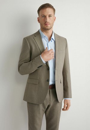 Mężczyzna w beżowym garniturze i jasnobłękitnej koszuli poprawiający marynarkę, stojący na jednolitym białym tle.