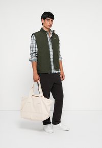 Gepatchte grüne Weste über einem blau-weiß karierten Hemd, schwarze Hose und weiße Sneaker. Hält eine große beigefarbene Canvas-Tasche.