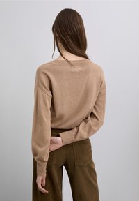 Suéter beige de punto con un corte relajado, cuello redondo y puños acanalados, combinado con pantalones de pana marrones. Detalle sutil de botones en la parte trasera.