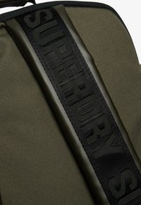 Zbliżenie na teksturowany materiał w kolorze oliwkowym z czarnym paskiem z taśmy, na którym widnieje wytłoczone logo "Superdry".