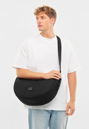 Čierna taška cez rameno vyrobená z odolnej tkaniny, s praktickým zaobleným tvarom, nastaviteľným popruhom a predným štítkom. Nosená cez telo.