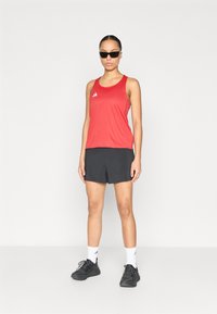 Red athletic tank top with a logo, paired with black shorts, white socks, and black sneakers. Model wears dark sunglasses.