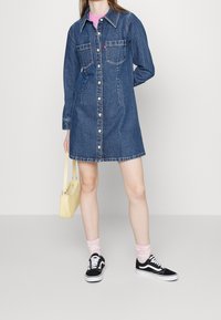 Sukienka koszulowa z denimu w ciemnym niebieskim kolorze, z długimi rękawami, dwoma przednimi kieszeniami, zapinana na guziki, o lekko rozkloszowanym kroju w literę A. Połączona z czarnymi trampkami.