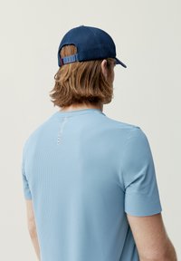 Světle modrá košile s krátkým rukávem a texturovaným povrchem, se bílým detailem na zadní části límce. V kombinaci s tmavě modrým síťovaným kloboukem.