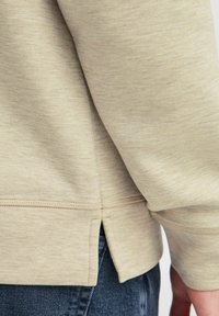Beige langermet skjorte med teksturert finish, som har sideslisser i kanten og synlige sømdetaljer. Kombinert med denimbukser.