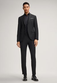 Completo nero su misura con pantaloni slim-fit e una camicia abbinata. I dettagli notevoli includono un fazzoletto bianco nella tasca e scarpe nere lucide.