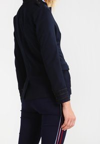 Giacca tailored blu navy con taglio strutturato, caratterizzata da bottoni decorativi e dettagli di cucitura intricata sui polsini. Tessuto dalla texture liscia.