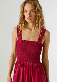 Νεαρή γυναίκα με κυματιστά ξανθά μαλλιά, που φοράει ένα κόκκινο αμάνικο φόρεμα με ελαστική μέση και βολάν στον λαιμό, στέκεται μπροστά σε μονόχρωμο φόντο.