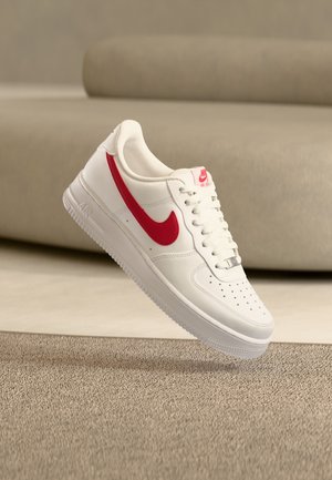 Biały sneaker Nike Air Force 1 z czerwonym logo Swoosh unoszący się nad beżowym dywanem i tłem sofy.