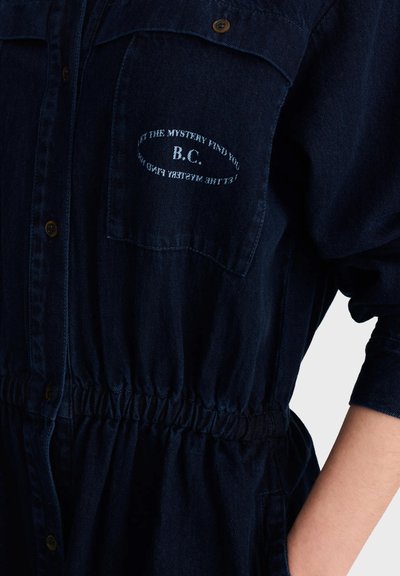 Chemise en jean bleu foncé avec taille élastique, fermeture à boutons, poche poitrine brodée avec "B.C." et texte circulaire "LET THE MYSTERY FIND YOU."