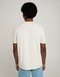 Ανοιχτό κρεμ μπλουζάκι κοντό μανίκι, από μαλακό βαμβακερό ύφασμα, με στρογγυλή τελείωση και άνετη εφαρμογή. Εμφανίζεται από την πλάτη, δείχνοντας τον απλό σχεδιασμό.