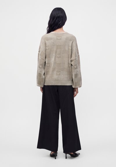 Femme aux longs cheveux foncés portant un pull beige à motifs, un pantalon noir à jambes larges et des chaussures noires à talons hauts, debout, de dos.
