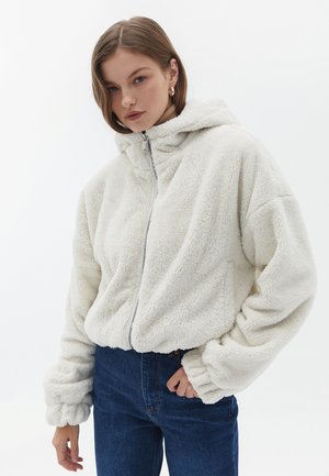Felpa con cappuccio in bianco sporco con chiusura a zip, vestibilità rilassata e polsini elasticizzati. Indossata sopra jeans blu scuro, con cappuccio.